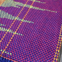 Die Abbildung zeigt den (Ausschnitt) des Schals, den eine Teilnehmerin beim TAB Web-Workshop gewebt hat. Der Schal hat die Farben blau, pink, gelb und apfelgrün gewebt und zeigt wellenartige oder strahlenförmige Muster.